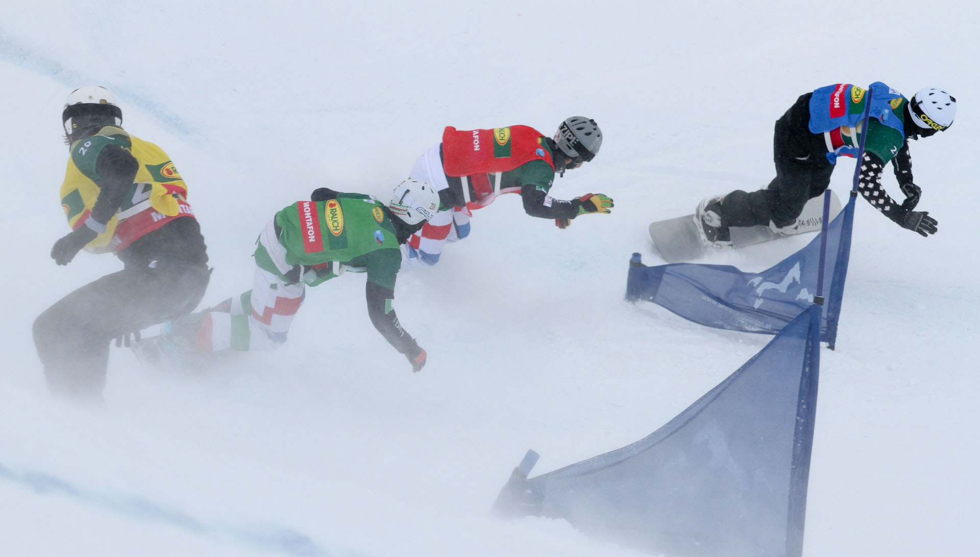 Four snowboard cross racers round a gate. 